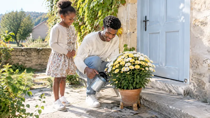 Jeune homme accroupi qui arrose au pied un chrysanthème blanc et jaune posé sur des cales en bois, sous le regard d'une fillette