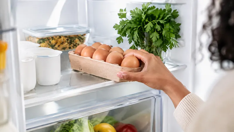 Gros plan sur une main qui range une boîte d'œufs frais dans un réfrigérateur, à côté de pots de yaourt blancs et d'un plat de restes
