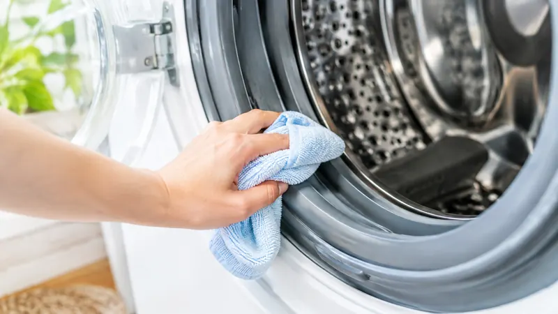 Main qui passe un chiffon microfibre bleu clair dans le pli en caoutchouc gris du joint d'un lave-linge ouvert