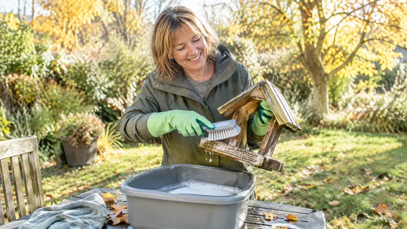 Une femme souriante en gants de jardinage brosse une mangeoire en bois au-dessus d'une bassine d'eau savonneuse, sur une table de jardin