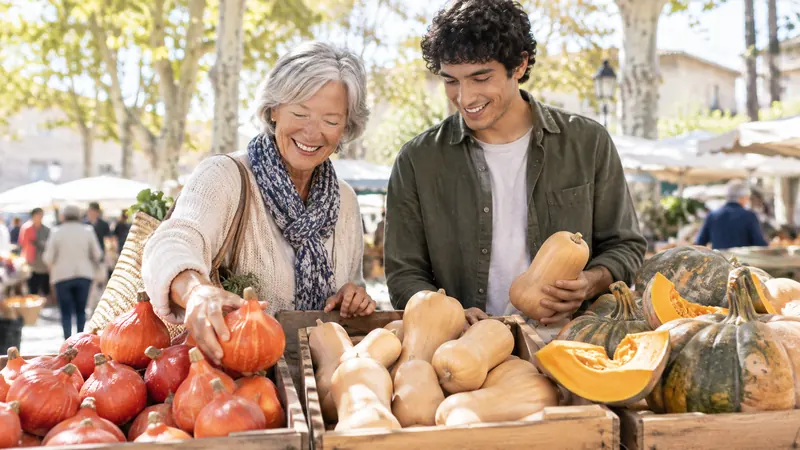Sur un étal de marché ensoleillé, une femme aux cheveux gris et un jeune homme choisissent des potimarrons, des butternuts et des tranches de courge