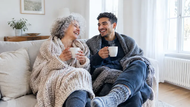 Une femme d'une soixantaine d'années et un jeune homme rient sur un canapé, enroulés dans des plaids, une tasse fumante à la main