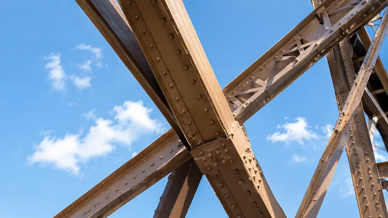 Contre-plongée sur des poutrelles rivetées peintes en brun, éclairées d'un côté par le soleil, sur fond de ciel bleu et de petits nuages